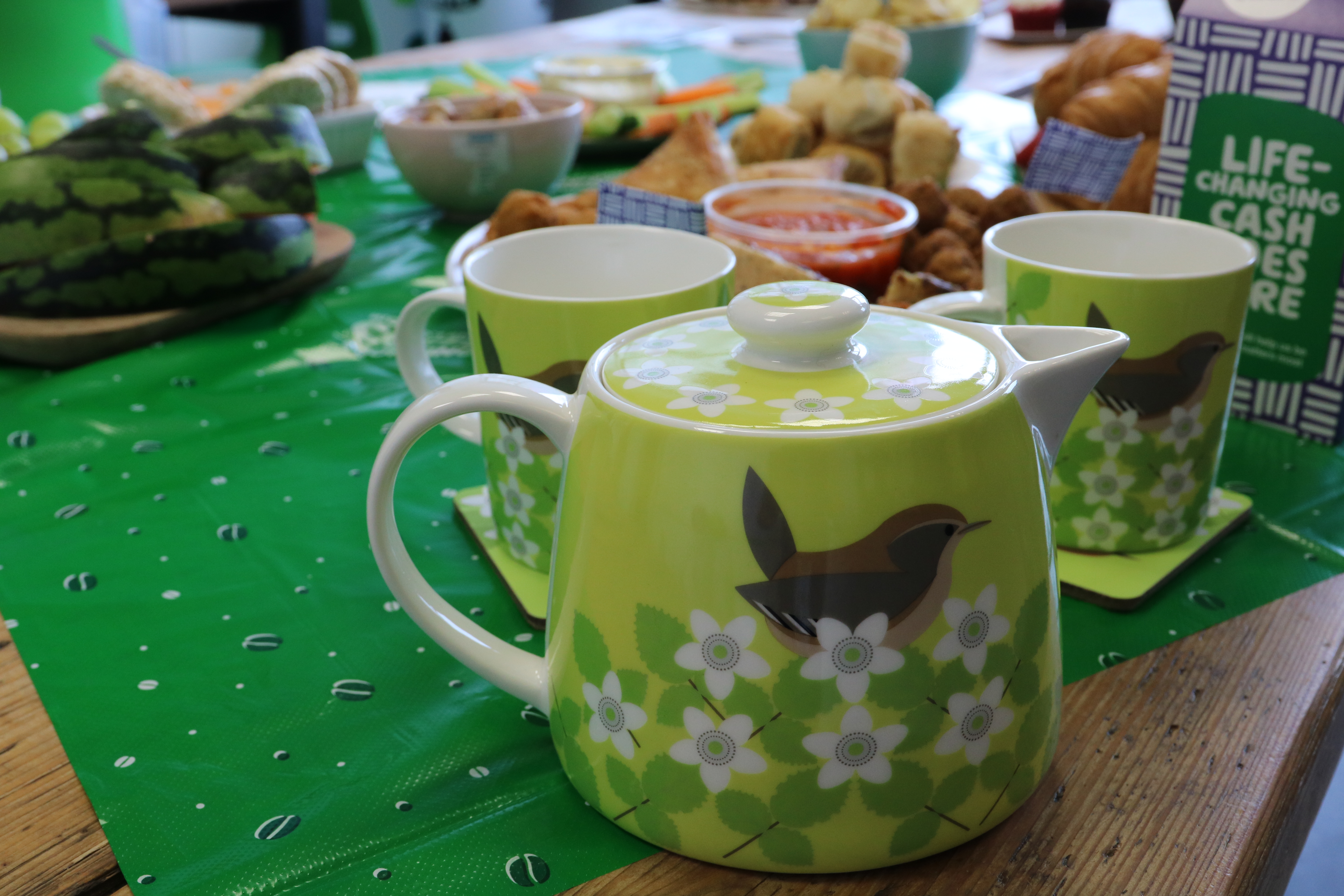 A green teapot on a table surrounded by cups and cakes.