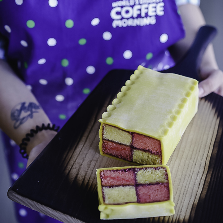 A cherry bakewell Battenberg cake on a chopping board, held by a woman with a Coffee Morning apron on.